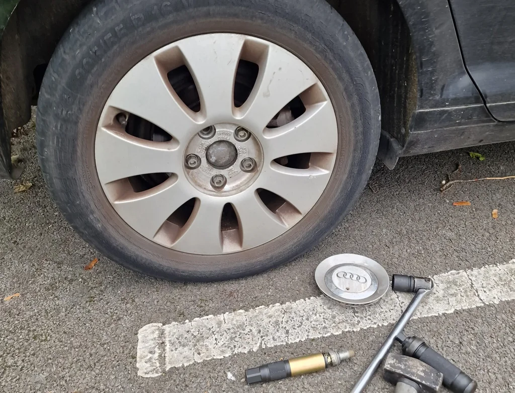 A flat car tyre with a removed hubcap, a tyre iron, and an inflation tool on the ground beside it, ready for mobile tyre fitting in Weymouth.