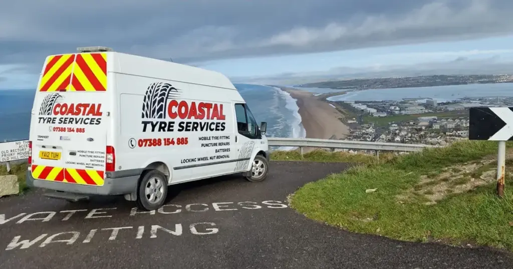 A service van from coastal tyre services parked by the roadside overlooking a coastal town and beach.