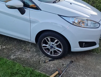 White car with a flat tire and locking wheel nut removal tools placed on the ground beside it in Weymouth.