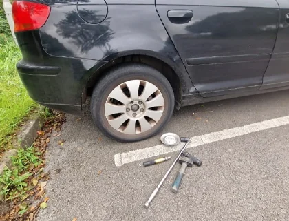 A car with a flat tire and tools, including a Locking Wheel Nut Removal kit from Weymouth, laid out on the ground for a tire change.